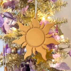 May include: A golden sun-shaped ornament with a textured surface, hanging from a white string. The ornament is part of a festive display, surrounded by purple flowers and a gold-colored tree.