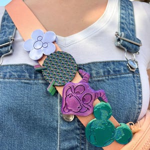 May include: A peach-colored strap adorned with colorful, decorative pins. The pins include a light purple flower, a green and pink patterned circle, a purple character, and a green Mickey Mouse-shaped design. The strap is attached to denim overalls.