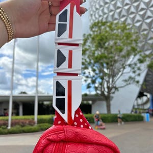 May include: Red faux leather fanny pack with white polka dots and a white and red monorail charm. The monorail charm is attached to the fanny pack with a pink strap.
