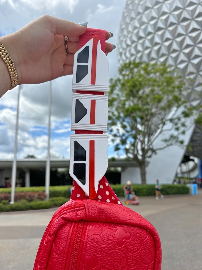 May include: Red faux leather fanny pack with white polka dots and a white and red monorail charm. The monorail charm is attached to the fanny pack with a pink strap.