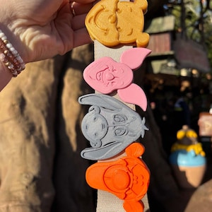 May include: A beige fabric strap with four colorful character charms attached. The charms are Winnie the Pooh, Piglet, Eeyore, and Tigger. The charms are made of 3D printed plastic.