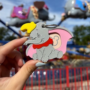 May include: A gray, pink, and yellow Dumbo keychain with a red bow. The keychain is shaped like Dumbo the elephant from the Disney movie.