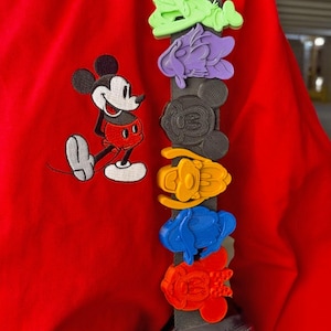 Könnte beinhalten: Rote Jacke mit weißem Streifen am Kragen und Mickey Mouse-Stickerei. Ein schwarzer Riemen mit bunten Mickey Mouse-förmigen Anhängern in Grün, Lila, Schwarz, Gelb, Blau und Rot.