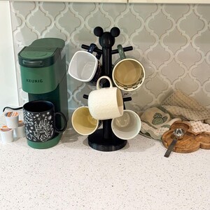 May include: A matte black mug tree with a Mickey Mouse ear topper holding six assorted ceramic mugs, positioned next to a forest green Keurig coffee maker, a small wooden Mickey-shaped tray, and a silver spoon on a white speckled countertop.