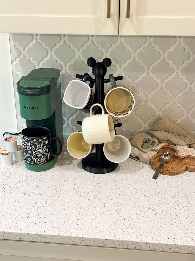May include: A matte black mug tree with a Mickey Mouse ear topper holding six assorted ceramic mugs, positioned next to a forest green Keurig coffee maker, a small wooden Mickey-shaped tray, and a silver spoon on a white speckled countertop.