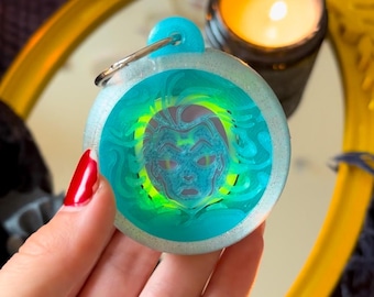 Könnte beinhalten: Ein blauer und grüner Schlüsselanhänger aus Harz mit einem wirbelnden Design und einem weißen Gesicht in der Mitte. Der Schlüsselanhänger hat einen silbernen Ring.