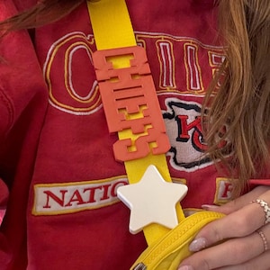 May include: A yellow and orange crossbody bag with a white star charm. The bag has a red strap with a red and yellow plastic charm that says "CHILL".