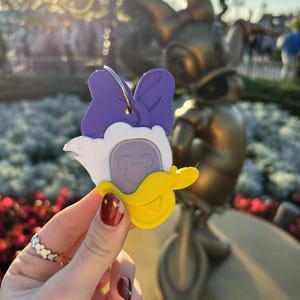 May include: Purple and white 3D printed keychain shaped like a duck with a bow. The duck has a yellow beak and a white face with a Mickey Mouse silhouette.