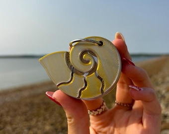 Op de afbeelding: Een gele en zilveren 3D-geprinte schelp sleutelhanger. De schelp is spiraalvormig met een getextureerd oppervlak.