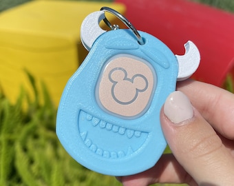 May include: A light blue keychain shaped like a monster with a white Mickey Mouse head on the front. The keychain has a white ring for attaching to a bag or keys.