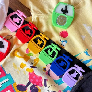 Könnte beinhalten: Eine Sammlung bunter Disney-Schlüsselanhänger in Pink, Rot, Gelb, Grün, Blau und Lila. Jeder Schlüsselanhänger zeigt ein Gondeldesign mit einer Charaktersilhouette. Die Schlüsselanhänger sind auf einem schwarzen Riemen angeordnet, mit einem gelben Shirt mit Disney-Figuren im Hintergrund.