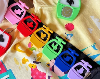 Könnte beinhalten: Eine Sammlung bunter Disney-Schlüsselanhänger in Pink, Rot, Gelb, Grün, Blau und Lila. Jeder Schlüsselanhänger zeigt ein Gondeldesign mit einer Charaktersilhouette. Die Schlüsselanhänger sind auf einem schwarzen Riemen angeordnet, mit einem gelben Shirt mit Disney-Figuren im Hintergrund.