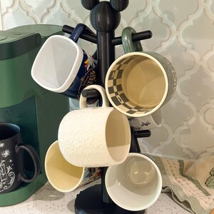 May include: A black mug tree stand with a mouse ear silhouette at the top, holding five ceramic mugs of various colors and patterns, including a white mug with a textured mouse head design and a dark blue mug with printed text.