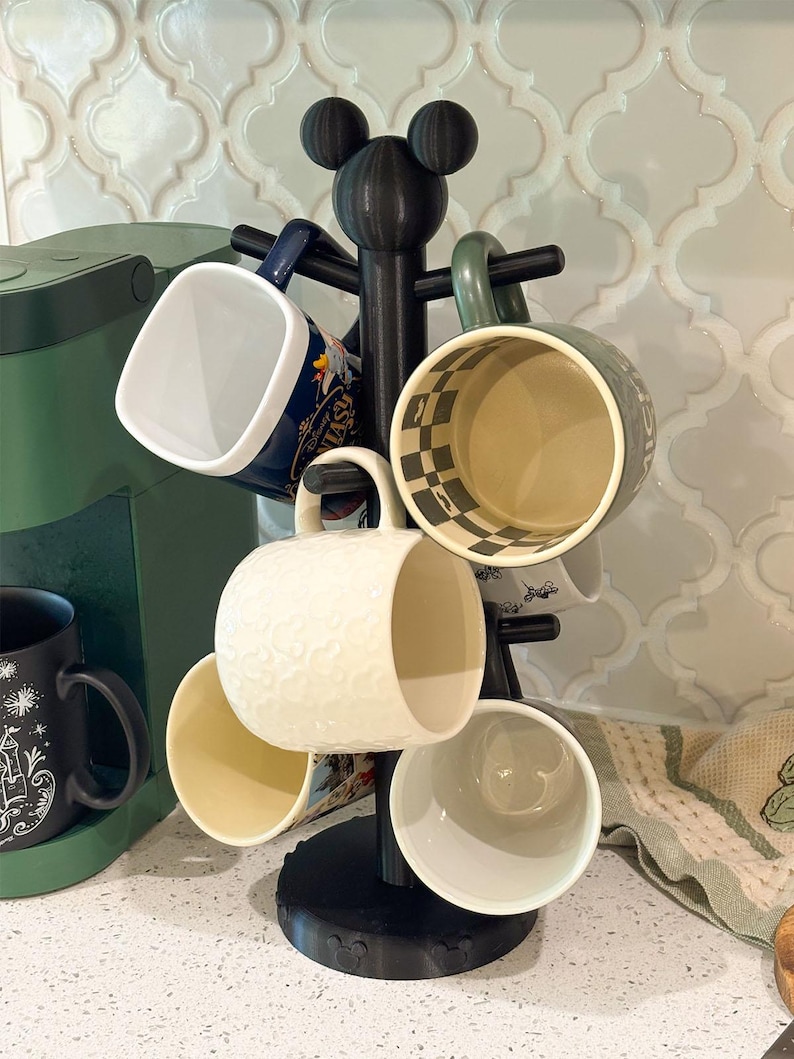 May include: A black mug tree stand with a mouse ear silhouette at the top, holding five ceramic mugs of various colors and patterns, including a white mug with a textured mouse head design and a dark blue mug with printed text.