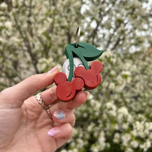 May include: A red and green cherry-shaped keychain with Mickey Mouse ears. The keychain is made of plastic and has a silver ring.