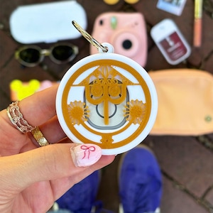 May include: A white and gold circular keychain with a decorative design. The keychain is held in front of a collection of accessories, including sunglasses, a pink camera, and lip gloss.