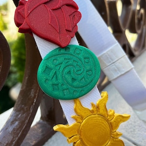 May include: A white headband adorned with colorful 3D-printed charms. The charms include a red rose, a green circular design, a gold sun, and a blue flower. The headband is set against a blurred outdoor background.