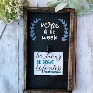 Puede incluir: Una pizarra negra con un marco de madera y una tarjeta blanca sujeta a ella. La pizarra dice "verse of the week" con un diseño de corona azul. La tarjeta blanca dice "be strong be brave be fearless Joshua 1:9 You are never alone".