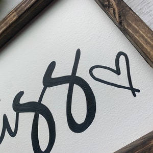 May include: White wooden sign with a dark brown frame. The sign features the word "bliss" in a black, cursive font, with a black heart symbol. The sign is a decorative item.