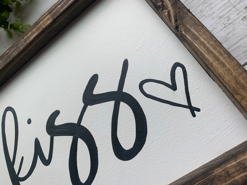 May include: White wooden sign with a dark brown frame. The sign features the word "bliss" in a black, cursive font, with a black heart symbol. The sign is a decorative item.