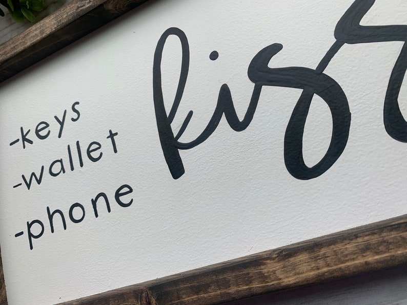 May include: A rectangular wooden sign with a white background and a dark brown frame. The sign features the word "first" in large, black cursive letters, along with a list of items: keys, wallet, and phone, in a smaller, black sans-serif font.
