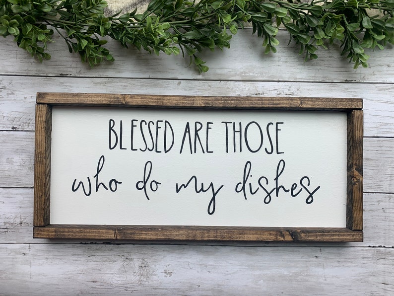 May include: A rectangular wooden sign with a white background and a dark brown frame. The sign features the phrase "BLESSED ARE THOSE who do my dishes" in black cursive lettering. Decorative greenery is visible above the sign.