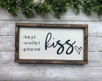 May include: A rectangular wooden sign with a white background and a dark brown frame. The sign features the words "-keys -wallet -phone" on the left and the word "kiss" with a heart symbol on the right.
