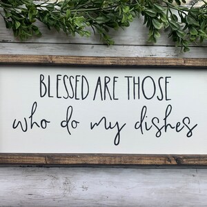 May include: A rectangular wooden sign with a white background and a dark brown frame. The sign reads "BLESSED ARE THOSE who do my dishes" in black script. Greenery is visible at the top.
