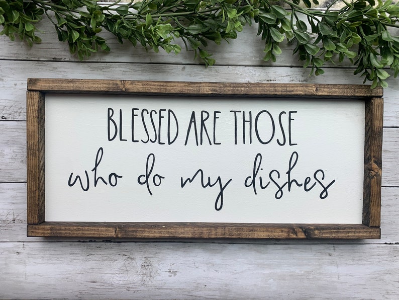 May include: A rectangular wooden sign with a white background and a dark brown frame. The sign reads "BLESSED ARE THOSE who do my dishes" in black script. Greenery is visible at the top.
