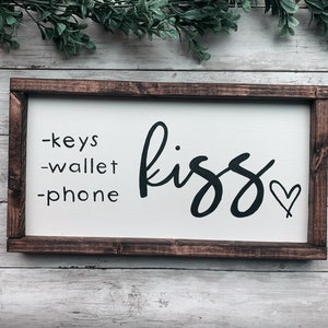 May include: A rectangular wooden sign with a white background and a dark brown frame. The sign features the words "-keys -wallet -phone" on the left and the word "kiss" with a heart symbol on the right in black script.