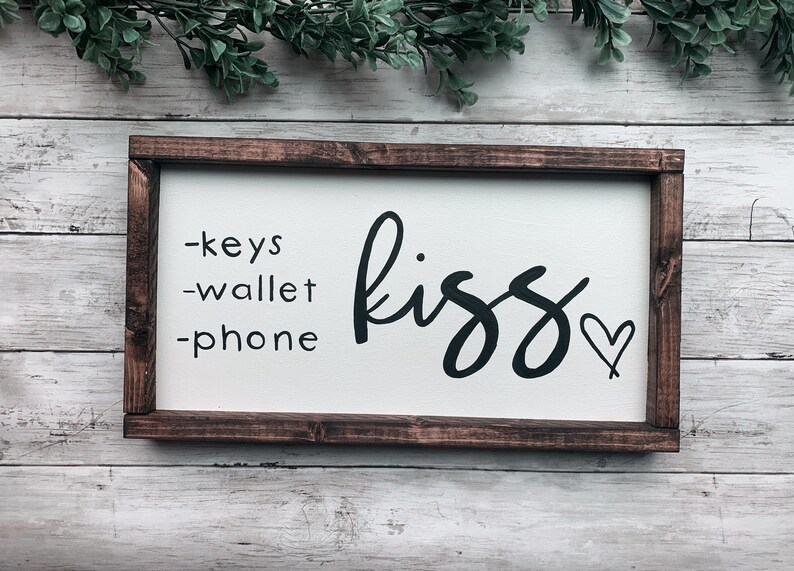 May include: A rectangular wooden sign with a white background and a dark brown frame. The sign features the words "-keys -wallet -phone" on the left and the word "kiss" with a heart symbol on the right in black script.