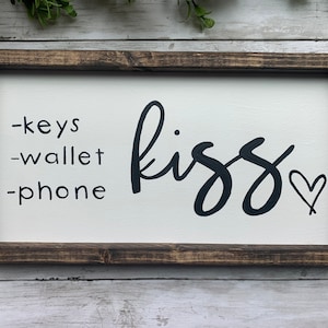 May include: A rectangular wooden sign with a white background and a dark brown frame. The sign features the words "-keys -wallet -phone" on the left and the word "kiss" with a heart symbol on the right.