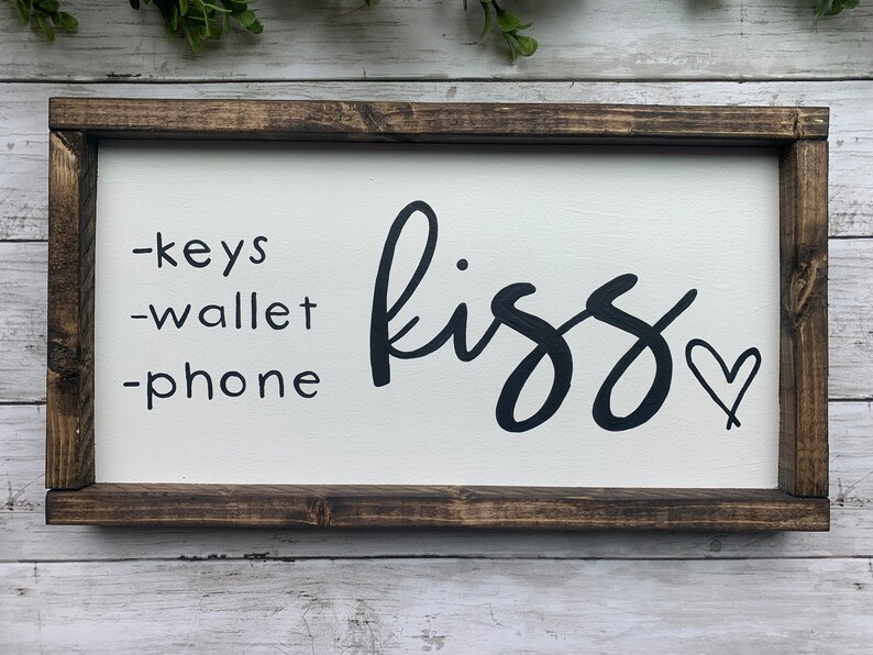 May include: A rectangular wooden sign with a white background and a dark brown frame. The sign features the words "-keys -wallet -phone" on the left and the word "kiss" with a heart symbol on the right.