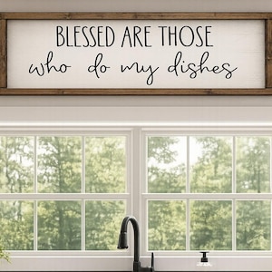 May include: A rectangular wooden sign with the text "BLESSED ARE THOSE who do my dishes" in black script. The sign is mounted above a window with a view of trees. The kitchen setting includes a sink and cabinets.