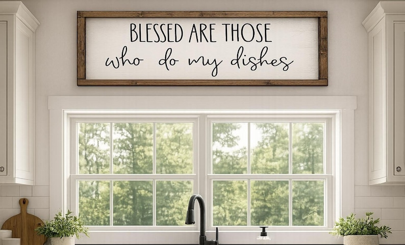 May include: A rectangular wooden sign with the text "BLESSED ARE THOSE who do my dishes" in black script. The sign is mounted above a window with a view of trees. The kitchen setting includes a sink and cabinets.