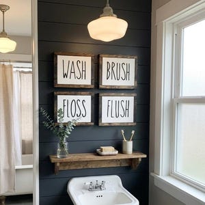 May include: Bathroom wall decor featuring four framed signs with the words "WASH", "BRUSH", "FLOSS", and "FLUSH" in black lettering. A wooden shelf holds a vase, soap, and toothbrushes. The wall is dark blue.