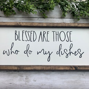 May include: A rectangular wooden sign with a white background and a dark brown frame. The sign features the phrase "BLESSED ARE THOSE who do my dishes" in black cursive lettering. Decorative greenery is visible at the top.