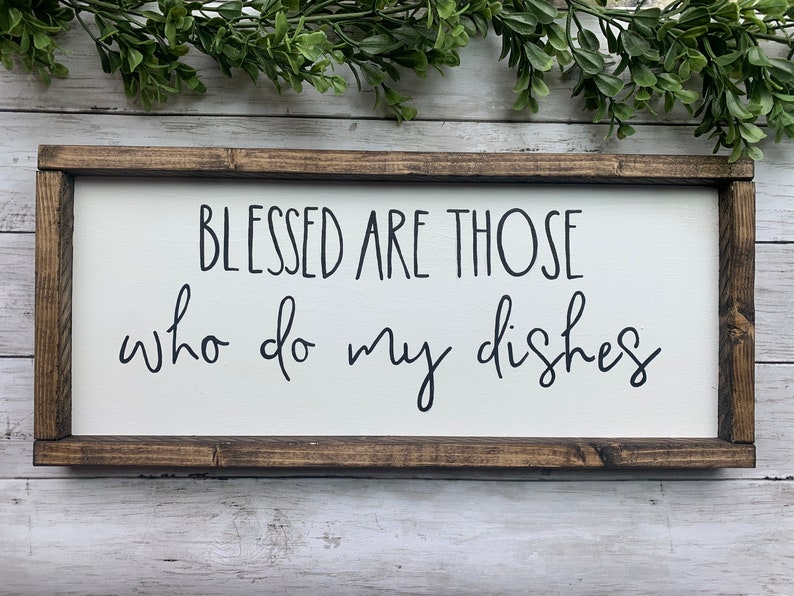 May include: A rectangular wooden sign with a white background and a dark brown frame. The sign features the phrase "BLESSED ARE THOSE who do my dishes" in black cursive lettering. Decorative greenery is visible at the top.