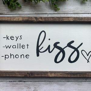 May include: A rectangular wooden sign with a white background and a dark brown frame. The sign has the words "-keys -wallet -phone" in black, and the word "kiss" in cursive, also in black, with a heart symbol.