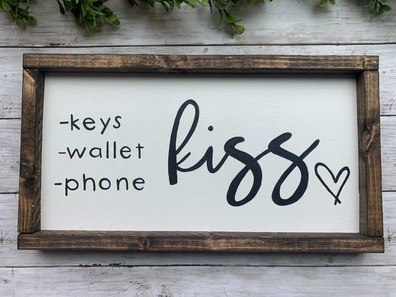 May include: A rectangular wooden sign with a white background and a dark brown frame. The sign has the words "-keys -wallet -phone" in black, and the word "kiss" in cursive, also in black, with a heart symbol.