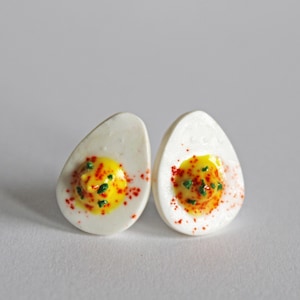May include: A pair of white ceramic earrings shaped like deviled eggs. The yolks are yellow with green chives and red paprika.