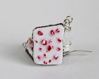 May include: A pair of white chocolate earrings with red candy pieces. The earrings are made of polymer clay and have silver wire hooks.