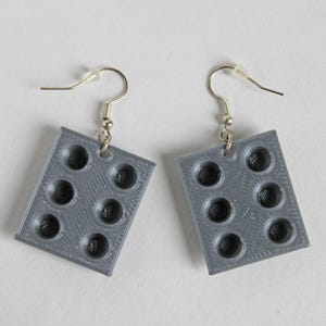 Peut inclure: Une paire de boucles d'oreilles carrées grises. Chaque boucle d'oreille présente un motif moulé ressemblant à un moule à muffins avec sept creux circulaires. Les boucles d'oreilles sont suspendues à des crochets d'oreille argentés.