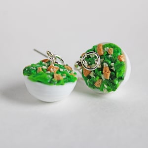 May include: A pair of white ceramic earrings shaped like bowls filled with green salad and croutons.