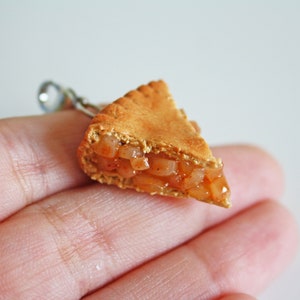 May include: A miniature apple pie slice charm, made of polymer clay, with a golden brown crust and a visible apple filling. The charm is held in a hand.