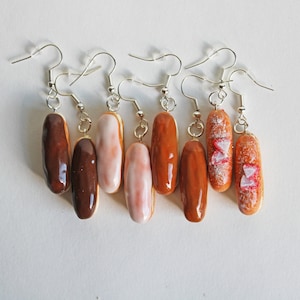 Könnte beinhalten: Ein Set mit sechs Paar Ohrringen, die Miniatur-Eclairs in verschiedenen Farben und Geschmacksrichtungen zeigen, darunter Schokolade, Vanille und Erdbeere.