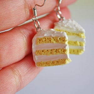 May include: A pair of dangle earrings featuring a miniature cake slice with white frosting and yellow cake layers.
