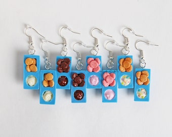 May include: Six pairs of blue and silver dangle earrings with miniature food-themed charms. Each charm features a blue square with two compartments. The top compartment has a variety of toppings, including sprinkles, chocolate chips, and small round cookies. The bottom compartment has a different type of ice cream, including vanilla, chocolate, strawberry, and a swirl of pink and white.