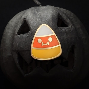 May include: A black jack-o'-lantern with a gold candy corn pin with a red, white, and yellow design. The candy corn has a smiling face.