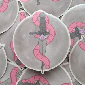 May include: A sticker featuring a pink worm wrapped around a silver dagger with a purple gemstone. The background is a gray circle with a galaxy-like pattern.